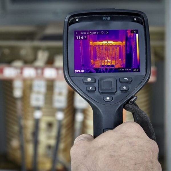 A FLIR E96 thermal camera held in front of an open electrical enclosure, its screen showing a live thermal image of the transformer windings with a spot reading of 114 degrees Fahrenheit on Asset C